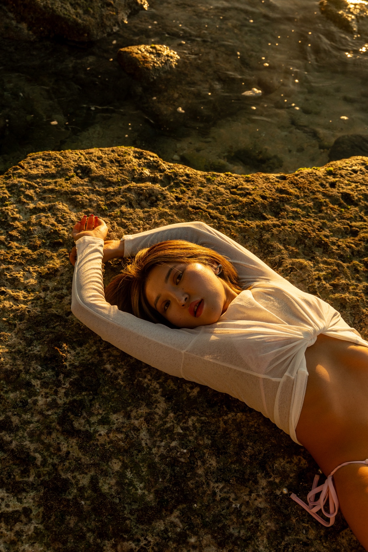Model in sheer white top resting on golden rock at sunset, Bali