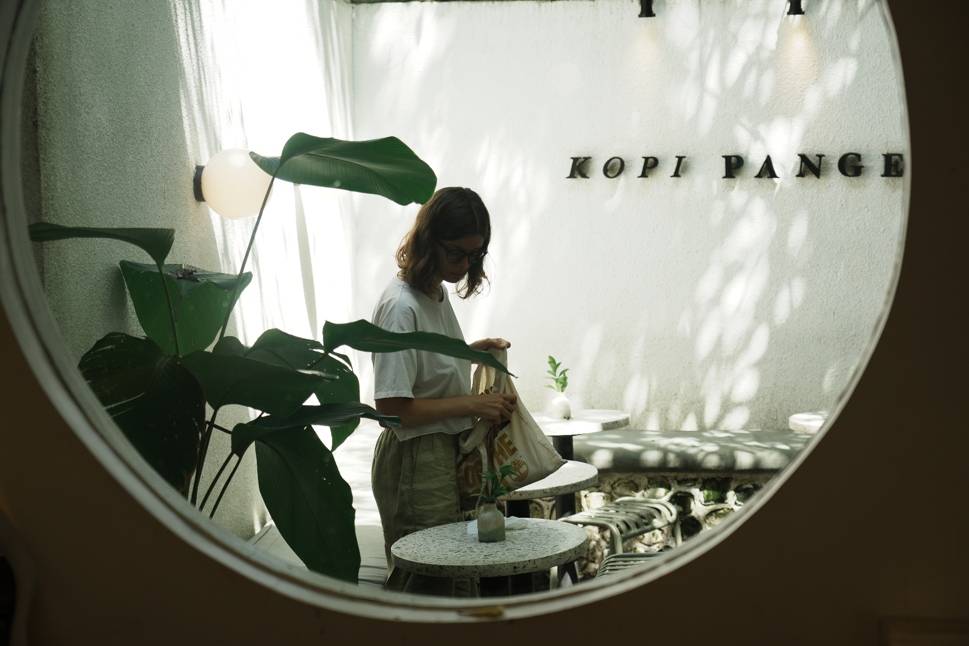 View through a circular porthole window into Kopi Pange — a guest browsing a tote bag among terrazzo tables and tropical leaves