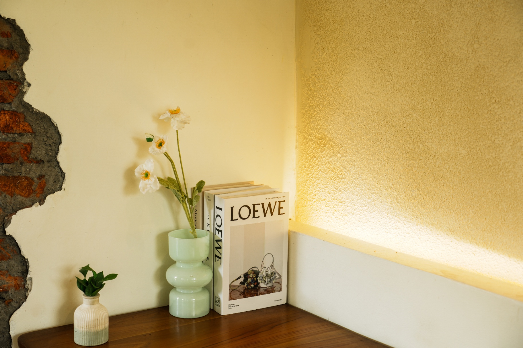 Still life in a quiet corner — Loewe monograph, jade glass vase with white poppies and warm rim light across the plaster wall